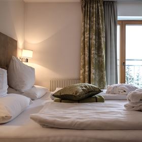 Ein gemütliches Schlafzimmer mit zwei Betten, weichen Kissen und frischen Handtüchern. Die Fenster sind mit grünen Vorhängen geschmückt und lassen viel Licht herein.