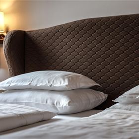 Ein gemütliches Schlafzimmer mit einem großen, gepolsterten Kopfteil und mehreren weißen Kissen. Im Hintergrund ist eine warm beleuchtete Lampe sichtbar.