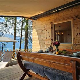 Ein gemütlicher Essbereich auf einer Veranda mit Blick auf den Wald und den See. Es gibt einen Holztisch und Liegen, die zum Entspannen einladen.