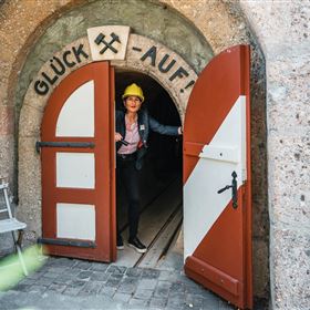 Un homme se tient devant une grande porte ouverte avec l'inscription « Bonne chance! ». Il porte un casque et regarde dans un étroit tunnel.