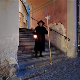 A person stands on a staircase with a lantern and a stick. The surroundings are illuminated at dusk and display historical architecture.