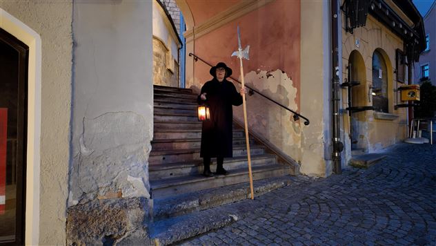 A person stands on a staircase with a lantern and a stick. The surroundings are illuminated at dusk and display historical architecture.