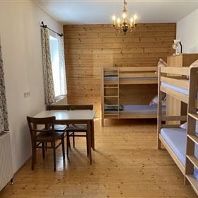A simple room with a wooden floor and a bunk bed. A table with chairs and a window with curtains are also available.