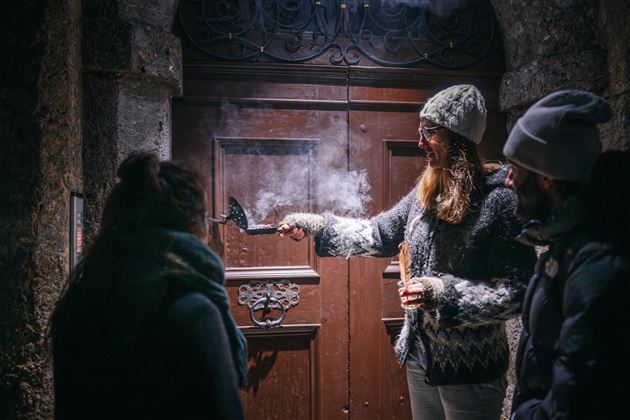 Eine Person steht vor einer Holztür und hält ein rauchendes Gerät. Zwei weitere Personen beobachten sie in einer winterlichen Umgebung.