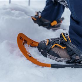 Ein Paar Schneeschuhe stehen im Schnee. Im Hintergrund sind die Beine eines Wanderers zu sehen, der bereit ist, in die verschneite Landschaft zu gehen.