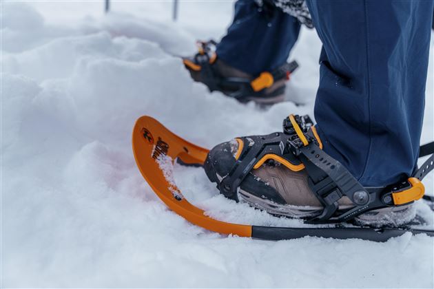 Ein Paar Schneeschuhe stehen im Schnee. Im Hintergrund sind die Beine eines Wanderers zu sehen, der bereit ist, in die verschneite Landschaft zu gehen.