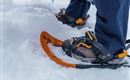 Ein Paar Schneeschuhe stehen im Schnee. Im Hintergrund sind die Beine eines Wanderers zu sehen, der bereit ist, in die verschneite Landschaft zu gehen.