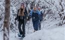 Drei Frauen wandern durch eine verschneite Waldlandschaft. Sie tragen Schneeschuhe und genießen die winterliche Umgebung.