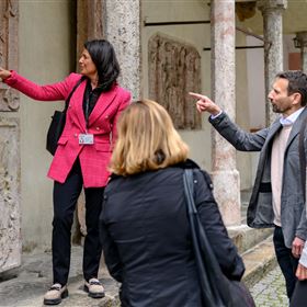 Eine Gruppe von Besuchern hört einer Führerin beim Erklären von Reliefs zu. Sie zeigen auf die Kunstwerke und stellen Fragen.