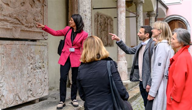 Eine Gruppe von Besuchern hört einer Führerin beim Erklären von Reliefs zu. Sie zeigen auf die Kunstwerke und stellen Fragen.