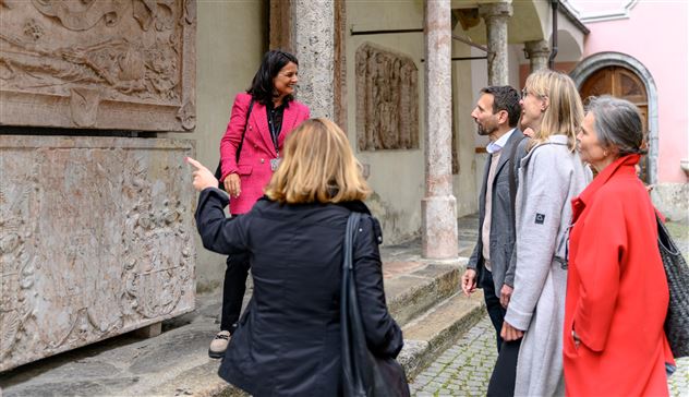 Eine Gruppe von fünf Personen steht vor einem historischen Gebäude. Eine Frau erklärt etwas und zeigt auf eine Wand mit Skulpturen.