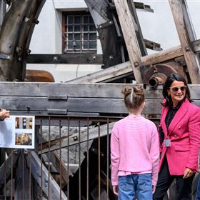 Un groupe de personnes regarde une grande structure en bois. Les participants écoutent attentivement une guide en veste rose.