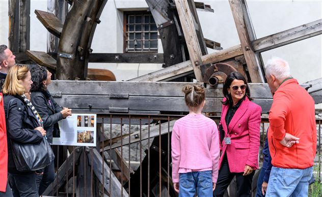 Eine Gruppe von Menschen betrachtet eine große Holzstruktur. Die Teilnehmer hören aufmerksam einer Führerin in pinker Jacke zu.