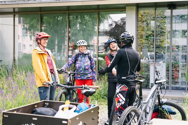 Four cyclists are conversing in front of a modern building. They are wearing helmets and casual clothing, surrounded by flowers.