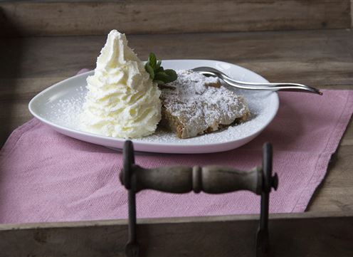 Un pezzo di torta con zucchero a velo e una grande porzione di panna montata su un piatto bianco. Il tutto è servito su un vassoio di legno con un tovagliolo rosa.