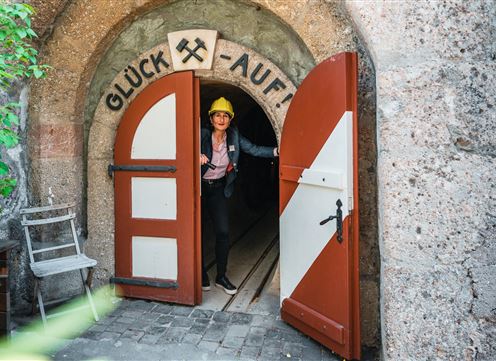 Een man staat voor een grote, open poort met de tekst "Geluk aan!". Hij draagt een helm en kijkt in een smalle tunnel.