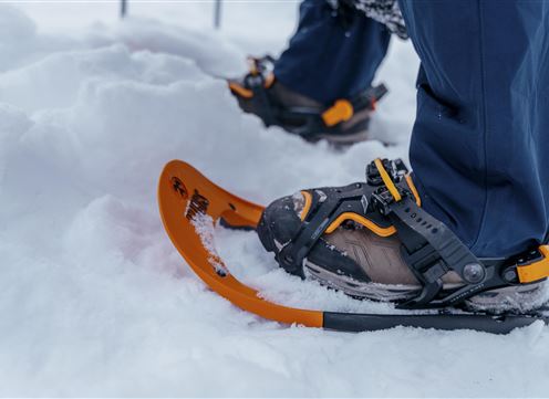 Ein Paar Schneeschuhe stehen im Schnee. Im Hintergrund sind die Beine eines Wanderers zu sehen, der bereit ist, in die verschneite Landschaft zu gehen.