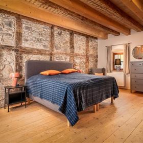 Ein stilvolles Schlafzimmer mit einem modernen Bett und orangefarbenen Kissen. Die Wände aus Stein und das Holzdesign verleihen dem Raum ein gemütliches Ambiente.