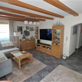 Ein helles Wohnzimmer mit Holzdeckenbalken und einer gemütlichen Couch. Auf der linken Seite befindet sich eine gepflegte Pflanze und ein TV-Möbelstück an der Wand.