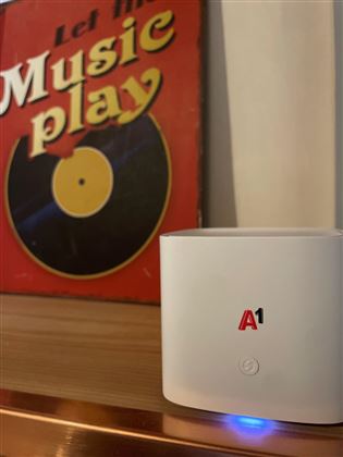 A modern streaming box is standing on a shelf. In the background, a colorful record cover can be seen, bearing the inscription "Let the Music Play."