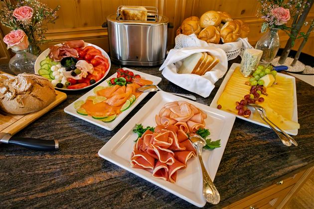 Un ricco buffet di colazione con vari piatti di affettati, frutta fresca e panini. La selezione comprende anche formaggi e prodotti da forno.