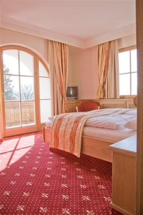 Una camera da letto luminosa con un grande letto e tende colorate. La luce entra dalle finestre e illumina il pavimento in moquette rossa.