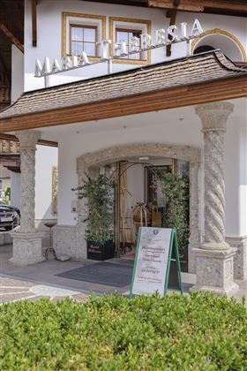 Un'accogliente area d'ingresso di un hotel con colonne e piante. Il cartello informa sui servizi e le offerte.