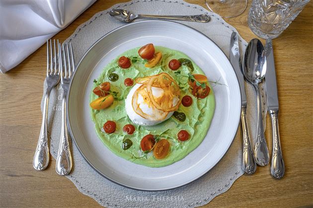 Un piatto gustoso su un piatto bianco con una salsa verde, pomodorini e un uovo morbido al centro. La tavola è elegantemente apparecchiata con posate e un bicchiere.