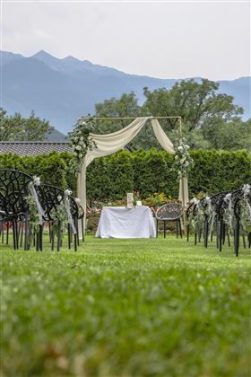 Un matrimonio romantico in giardino con un palcoscenico decorato in modo festoso e sedie su un prato verde. Sullo sfondo si possono vedere alberi e montagne.