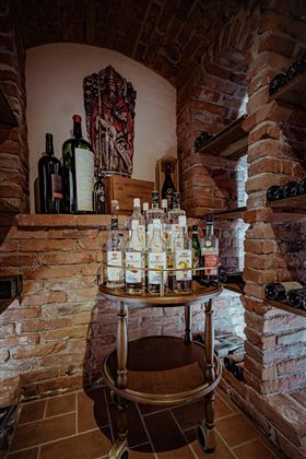 Un'ambiente vinicolo rustico con pareti in mattoni e un tavolo rotondo decorato con varie bevande. Sullo sfondo ci sono bottiglie e casse di vino sugli scaffali.