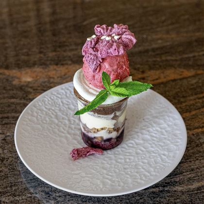 Un delizioso dessert a strati in un bicchiere con un ripieno cremoso, bacche e una pallina di gelato rosso sopra. È decorato con una foglia di menta e fiori commestibili.