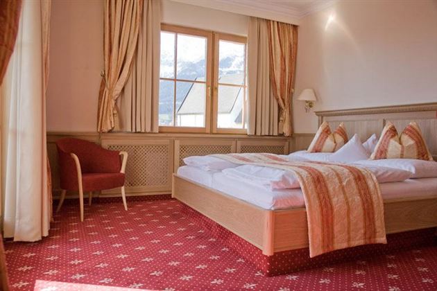 Una stanza d'hotel accogliente con un grande letto e cuscini eleganti. Grandi finestre offrono molta luce naturale e una vista sulle montagne.