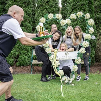 Una scena divertente con diverse donne che posano in una cornice a forma di cuore realizzata con fiori. Un uomo porta una coppa d'oro su un vassoio e crea sorpresa.