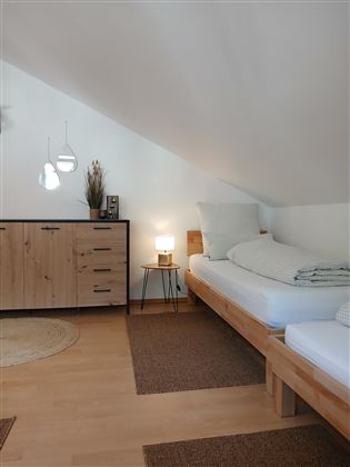 A cozy bedroom with two beds and a modern wooden furniture piece. The walls are light, and there is warm lighting.