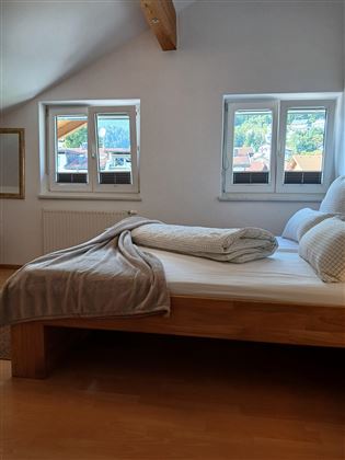 A cozy bedroom with a large wooden bed and two pillows. The windows let in daylight and offer a view outside.