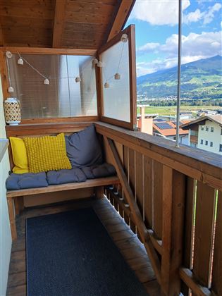 A cozy balcony with a seating area and cushions. In the background, a picturesque landscape and blue sky can be seen.