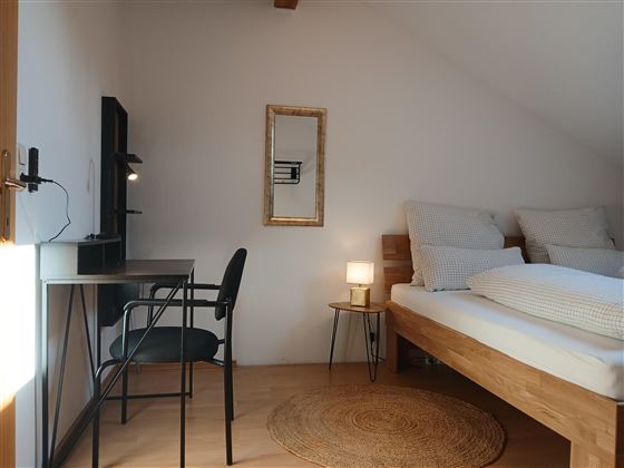 A simple bedroom with a cozy double bed and a desk. The room has a modern style and is well-lit.