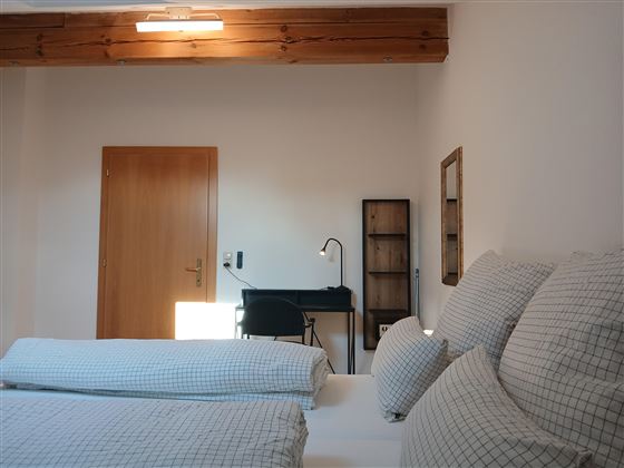 A bright room with two single beds and striped pillows. On the other side, there is a desk with a chair and a wall shelf.