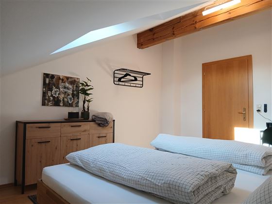 A bright room with two beds and wooden furniture. A picture hangs on the wall and there is a small wardrobe.