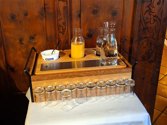 A tray with several glasses, a glass carafe with water, and one with orange juice. In the background, a wood-paneled wall is visible.
