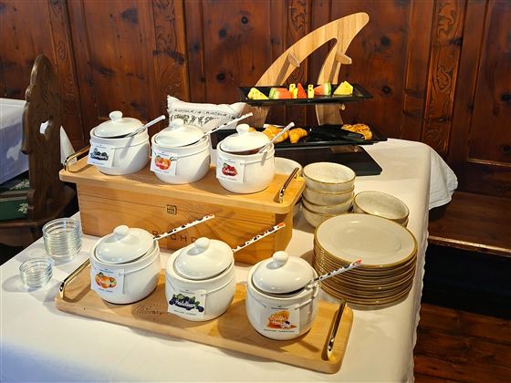 A pretty buffet with various teapots and an assortment of plates. In the background, fresh fruit and a decorative wooden sculpture can be seen.