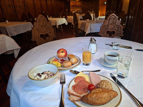 A laid table with various breakfast dishes, including fruit, yogurt, sausage, and rolls. The room has a cozy atmosphere with wooden furniture and white tablecloths.
