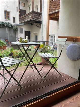 A small balcony with a table and two chairs. In the background, you can see a green garden and a calm courtyard atmosphere.