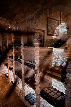 Una cantina con muri di mattoni e scaffali pieni di bottiglie di vino. La luce filtra da una finestra creando un'atmosfera accogliente.