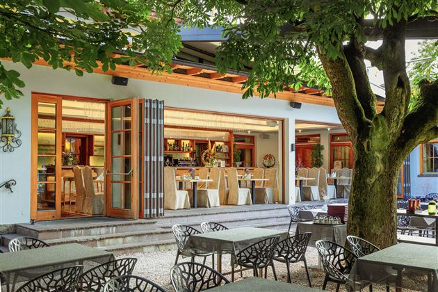 Un ristorante accogliente con porte aperte e tavoli all'aperto. Grandi alberi offrono ombra e creano un'atmosfera invitante.