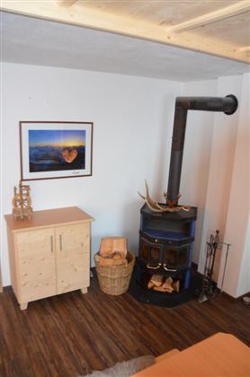 Een gezellige ruimte met een houtkachel in de hoek. Aan de muur hangt een afbeelding van een landschap, naast de kachel staat een mand met hout.