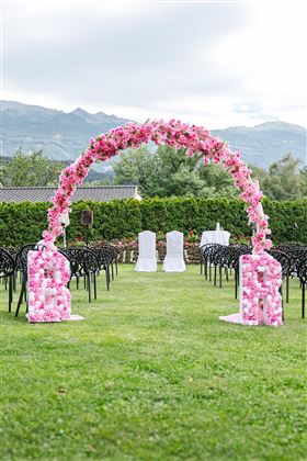 Un arco nuziale decorato con belle fiori rosa si trova su un prato verde. Davanti all'arco sono stati sistemati sedie per gli ospiti.