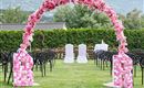 Un arco nuziale decorato con belle fiori rosa si trova su un prato verde. Davanti all'arco sono stati sistemati sedie per gli ospiti.