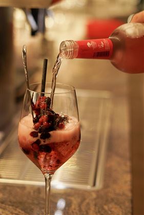 Un bicchiere da cocktail è riempito con una bevanda rosa e bacche fresche. Sullo sfondo si vede un bar sfocato.