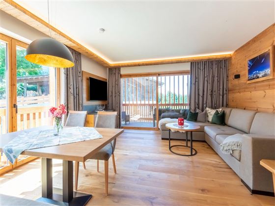 A bright and modern living room with large windows and wooden elements. The space is cozily furnished, with a dining table, a sofa set, and decorative plants.